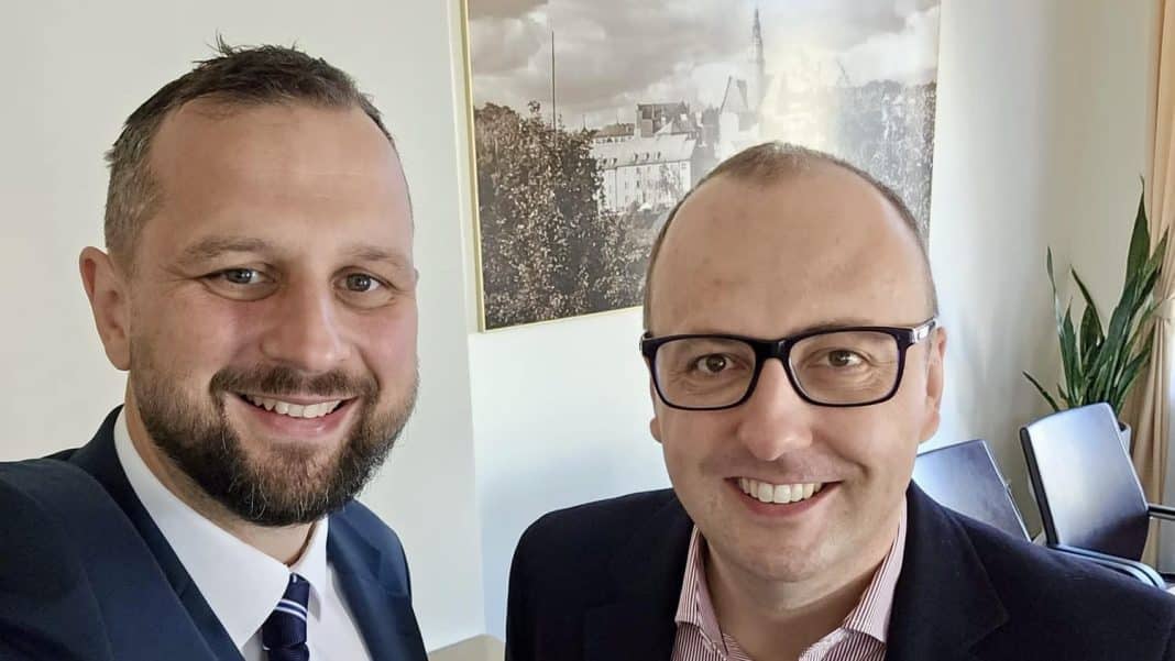 Dwa mężczyźni w biurze, uśmiechnięci, na tle obrazu miasta na ścianie.