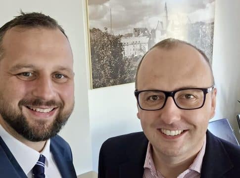 Dwa mężczyźni w biurze, uśmiechnięci, na tle obrazu miasta na ścianie.