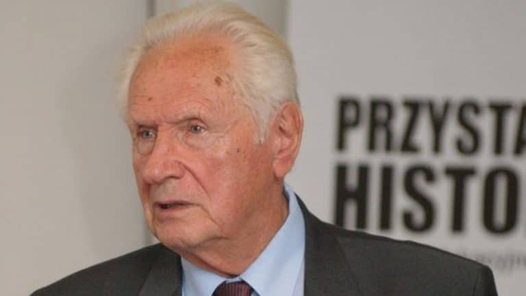 Starszy mężczyzna w garniturze na tle tablicy z napisem "Przystanek Historia".