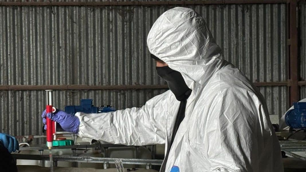 Osoba w ochronnym kombinezonie i masce pracuje w laboratorium na tle metalowych ścian.