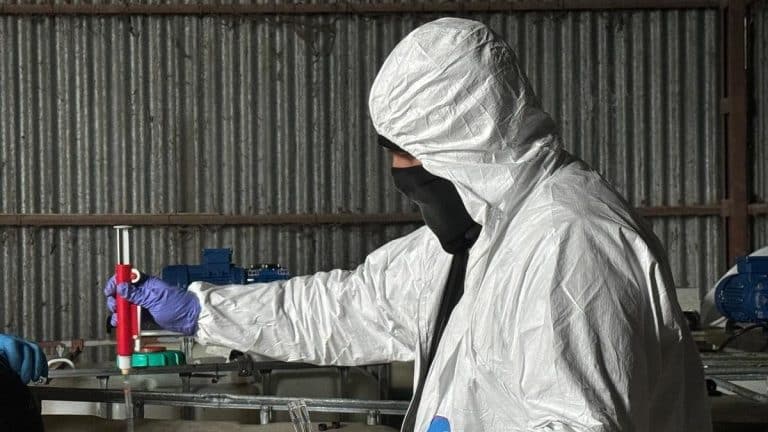 Osoba w ochronnym kombinezonie i masce pracuje w laboratorium na tle metalowych ścian.