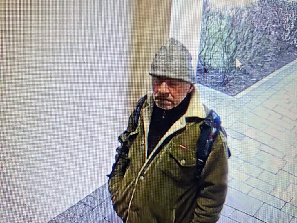 Mężczyzna w czapce i kurtce na monitoringu, patrzy w kamerę, na tle klatki schodowej.