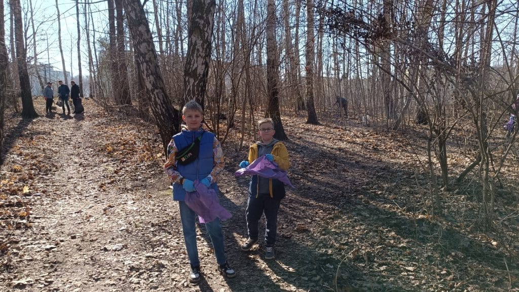 Dzieci z workami na śmieci na leśnej ścieżce podczas sprzątania lasu w słoneczny dzień.