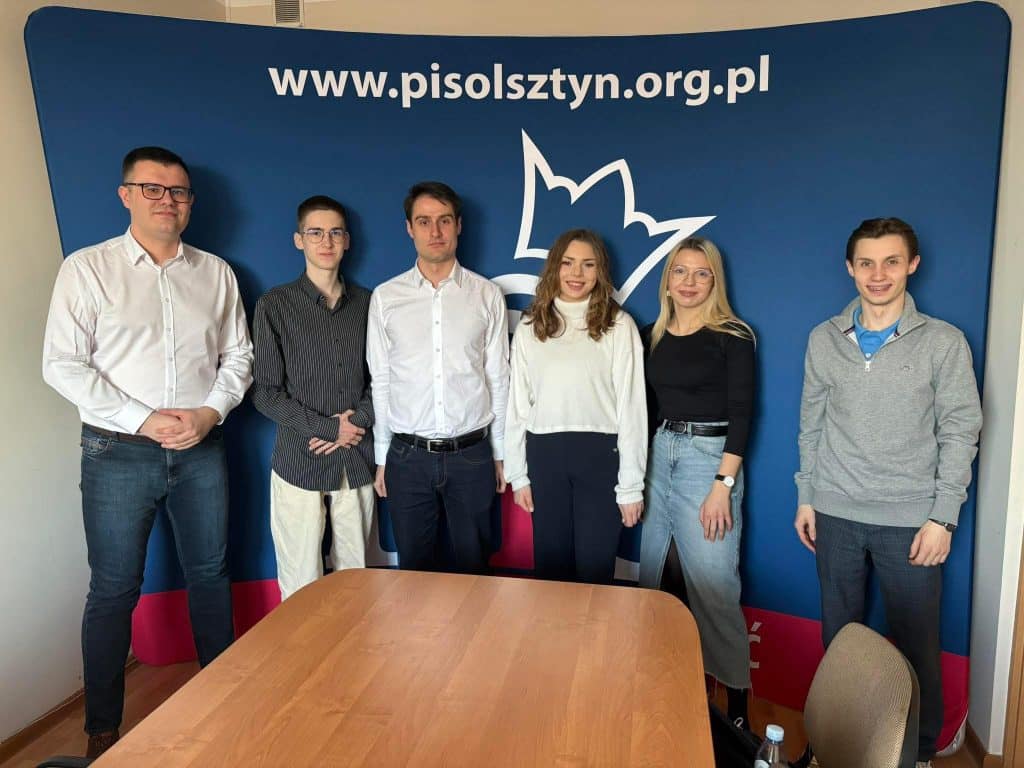 Grupa siedmiu os&oacute;b stojących przed logo organizacji na tle niebieskiej ściany.