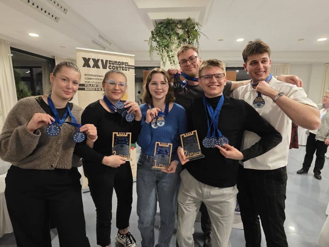 Grupa student&oacute;w UWM z medalami i trofeami na tle plakatu konkursu sensorycznego.