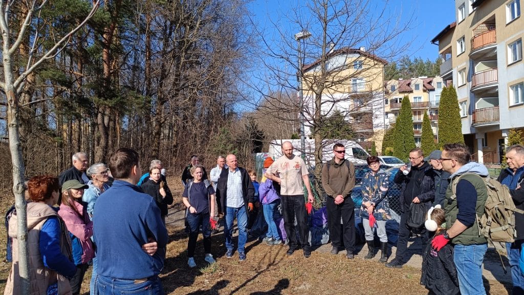 Grupa mieszkańc&oacute;w i uczestnik&oacute;w akcji sprzątania w lesie, w tle budynki i drzewa, słoneczna pogoda.