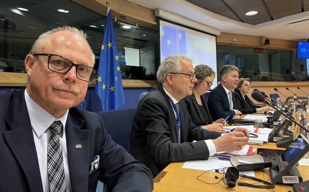 Zdjęcie przedstawia grupę osób podczas posiedzenia w sali konferencyjnej z flagami UE w tle.