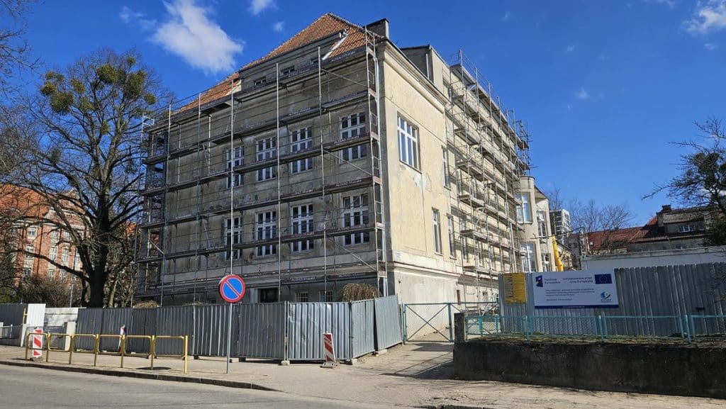 Budynek w trakcie remontu, otoczony rusztowaniami i ogrodzeniem, na tle nieba z chmurami.