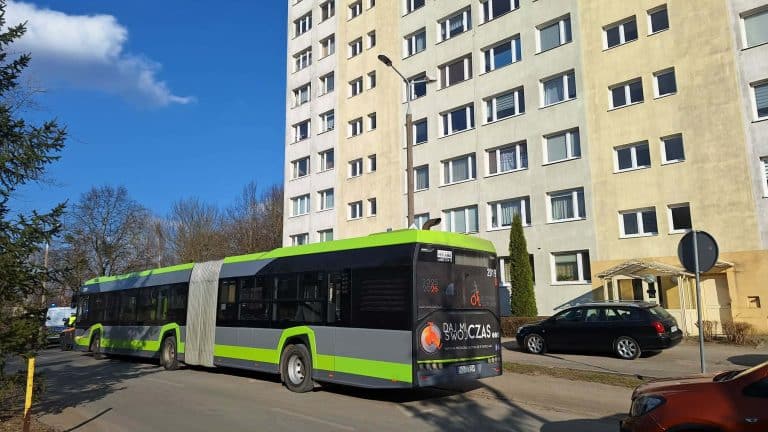 Miejski autobus na ulicy przed blokiem, w tle budynek mieszkalny, słoneczny dzień.