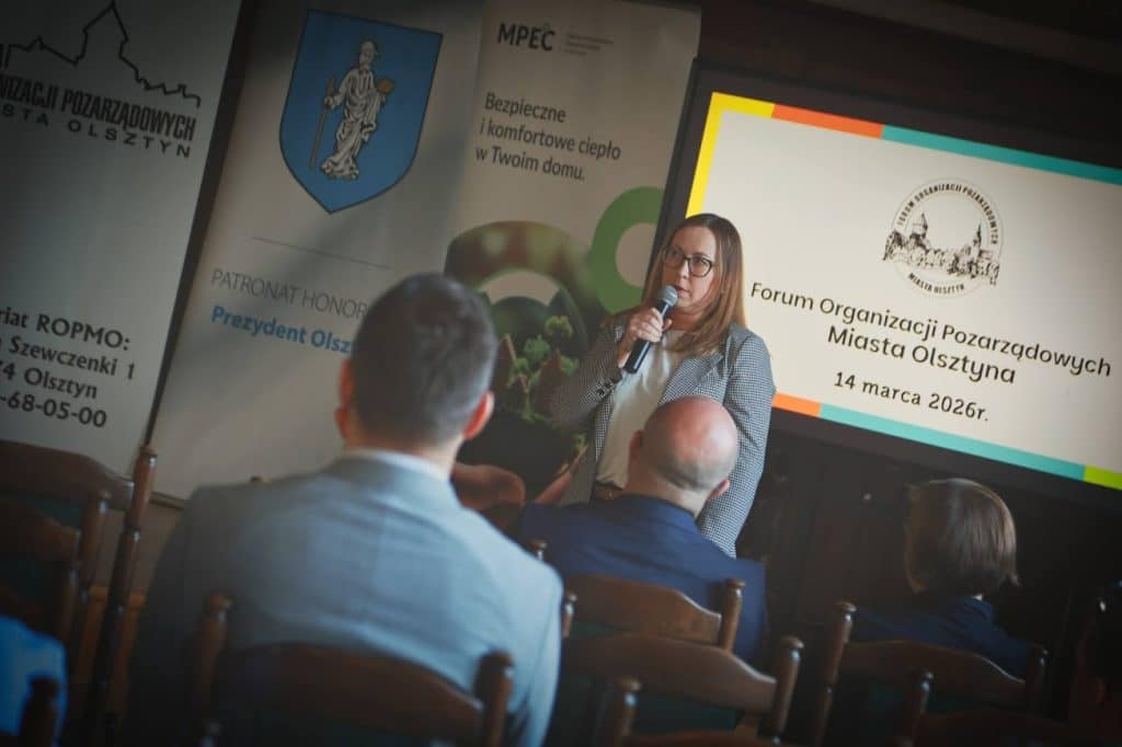 Kobieta przemawia do publiczności podczas spotkania organizacji społecznych w Olsztynie, za nią ekran z informacją o wyborze rady.