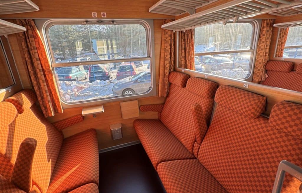Wnętrze pociągu z czerwono-pomarańczowymi tapicerkami i dużymi oknami, przez kt&oacute;re widać śnieg i samochody na zewnątrz.
