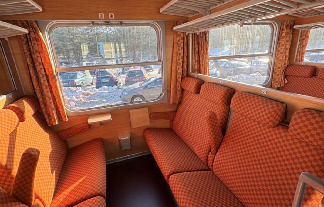 Wnętrze pociągu z czerwono-pomarańczowymi tapicerkami i dużymi oknami, przez kt&oacute;re widać śnieg i samochody na zewnątrz.