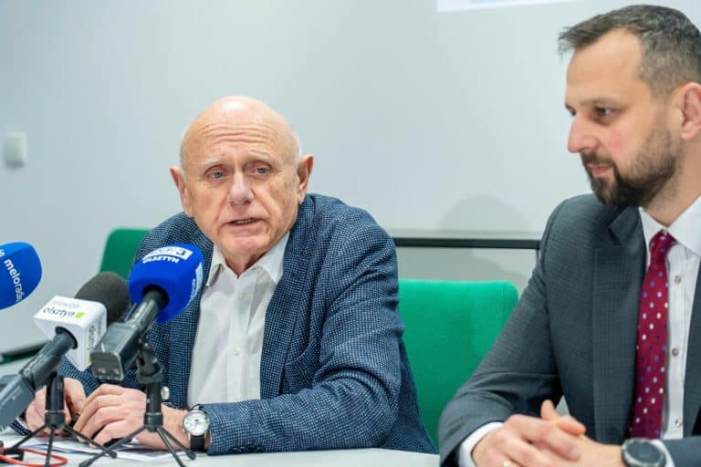 Dw&oacute;ch mężczyzn podczas konferencji prasowej, jeden m&oacute;wi do mikrofon&oacute;w, drugi słucha.
