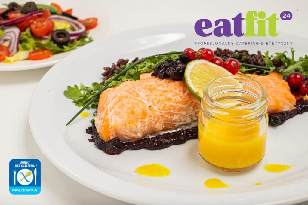 Danie z łososiem, warzywami i owocami na białym talerzu, obok mały słoik z ż&oacute;łtym sosem.