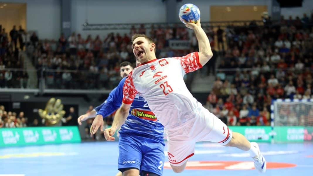 Piłkarz ręczny w białej koszulce z czerwonymi elementami, skaczący z piłką w hali sportowej, za nim tłum widz&oacute;w.