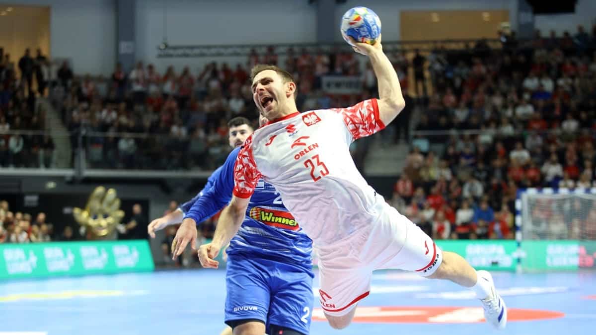 Piłkarz ręczny w białej koszulce z czerwonymi elementami, skaczący z piłką w hali sportowej, za nim tłum widzów.