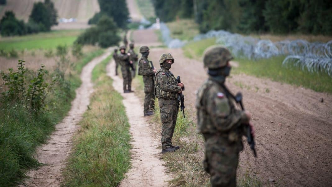 Grupa żołnierzy patrolujących granicę polsko-białoruską w terenie z drutem kolczastym w tle.