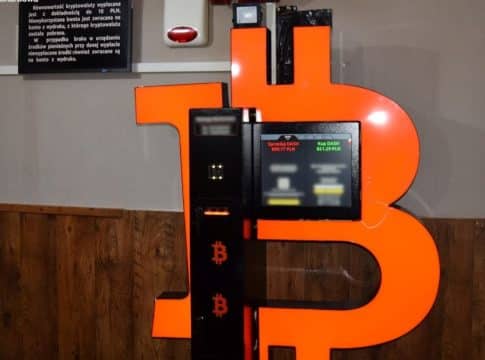 Bitomat z dużą literą B i ekranem wyświetlającym kursy kryptowalut, ustawiony w pomieszczeniu z drewnianą podłogą.