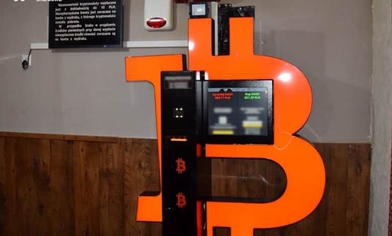 Bitomat z dużą literą B i ekranem wyświetlającym kursy kryptowalut, ustawiony w pomieszczeniu z drewnianą podłogą.