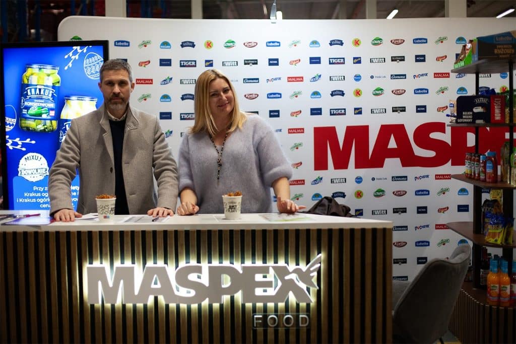 Stojący przy stoisku przedstawiciele branży spożywczej podczas targ&oacute;w w Gdańsku, za nimi logo i banery wystawc&oacute;w.