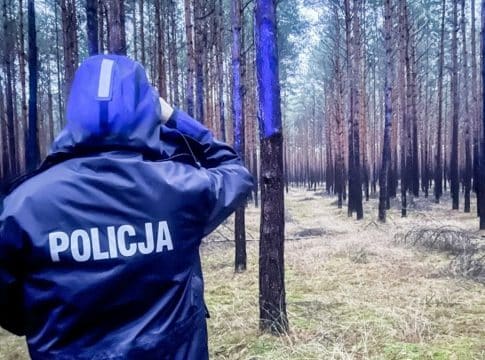 Policjant w lesie podczas poszukiwań zaginionej 17-latki, ubrany w kurtkę z napisem 'Policja'.