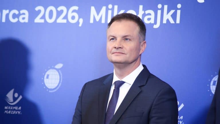 Zdjęcie mężczyzny w garniturze na tle niebieskiej ściany z napisami.