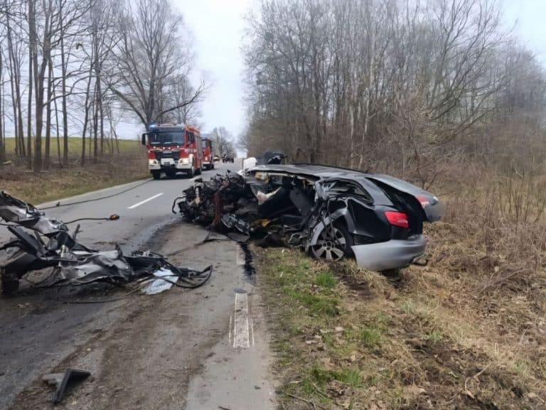 Rozbity samoch&oacute;d na poboczu po wypadku na drodze, w tle straż pożarna i zniszczone auto.