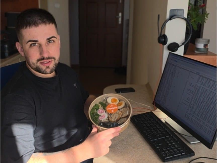 Mężczyzna trzyma miskę z jedzeniem w biurze, obok komputer i słuchawki na mikrofonie.