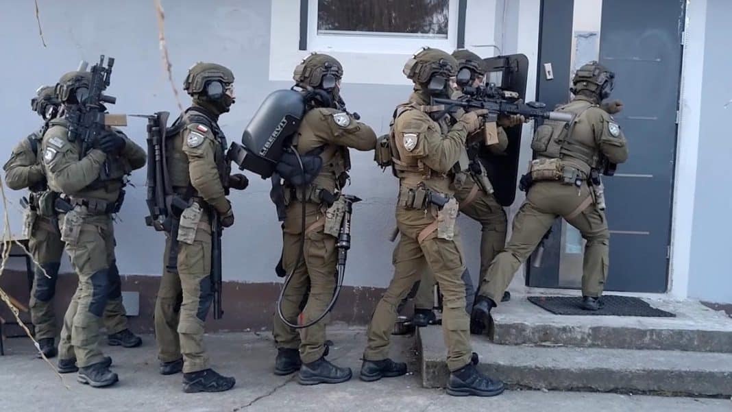 Grupa funkcjonariuszy w pełnym umundurowaniu i z bronią podczas akcji policyjnej przy drzwiach budynku.