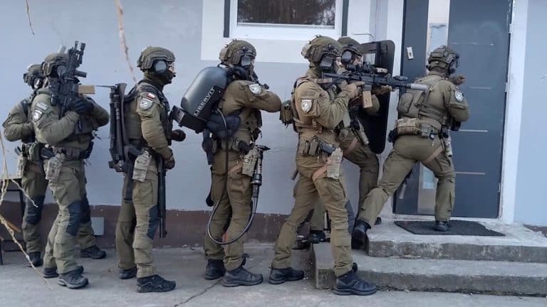 Grupa funkcjonariuszy w pełnym umundurowaniu i z bronią podczas akcji policyjnej przy drzwiach budynku.