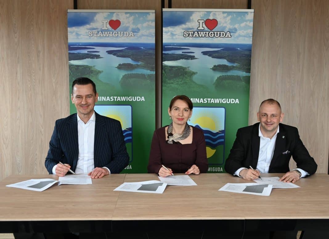 Trzy osoby siedzą przy stole, podpisując dokumenty, z dwoma plakatami w tle z napisem