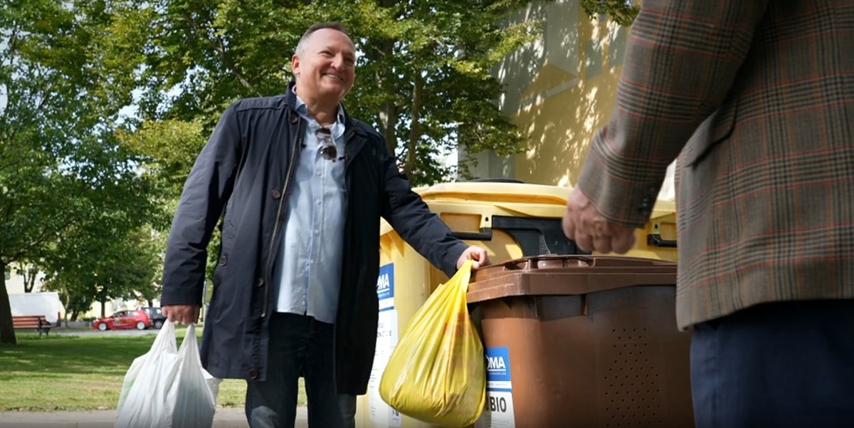 Mężczyzna wrzuca bioodpady do pojemnika na tle zielonego parku, uśmiecha się podczas segregacji odpadów.