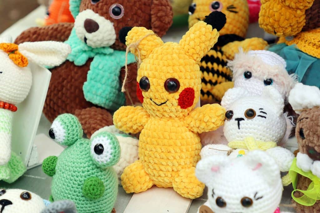 Ręcznie robione pluszowe zabawki, gł&oacute;wnie zwierzątka, na tle innych podobnych przedmiot&oacute;w.