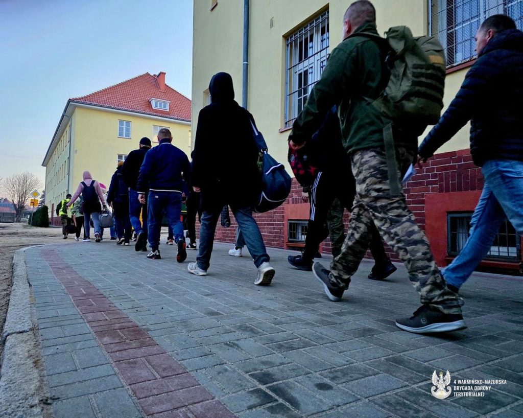 Grupa ludzi wchodząca do budynku wojskowego w Braniewie, wczesny poranek.