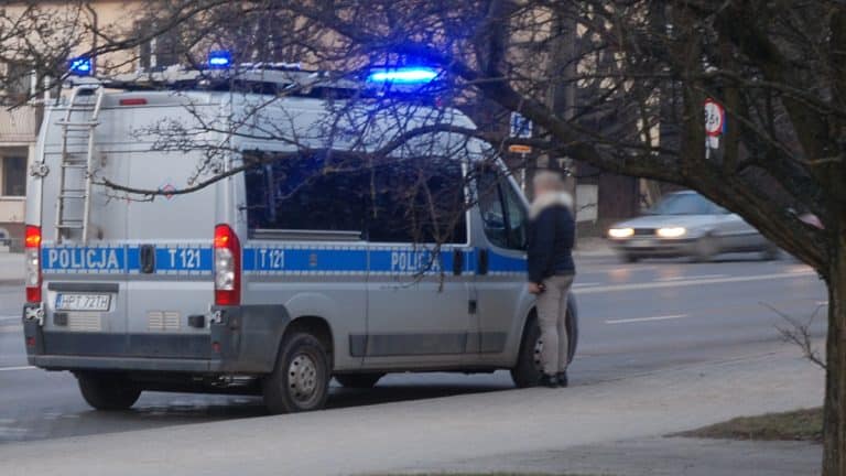 Mężczyzna stojący obok policyjnego radiowozu na przystanku, w tle ruchliwa ulica, widok z boku.