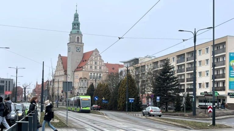Ulica w Olsztynie z tramwajem, samochodami i pieszymi, w tle zabytkowy ratusz i nowoczesne budynki.