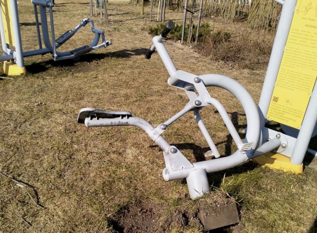 Zniszczona rowerowa siłownia na zewnątrz, przewr&oacute;cona i uszkodzona na trawniku.