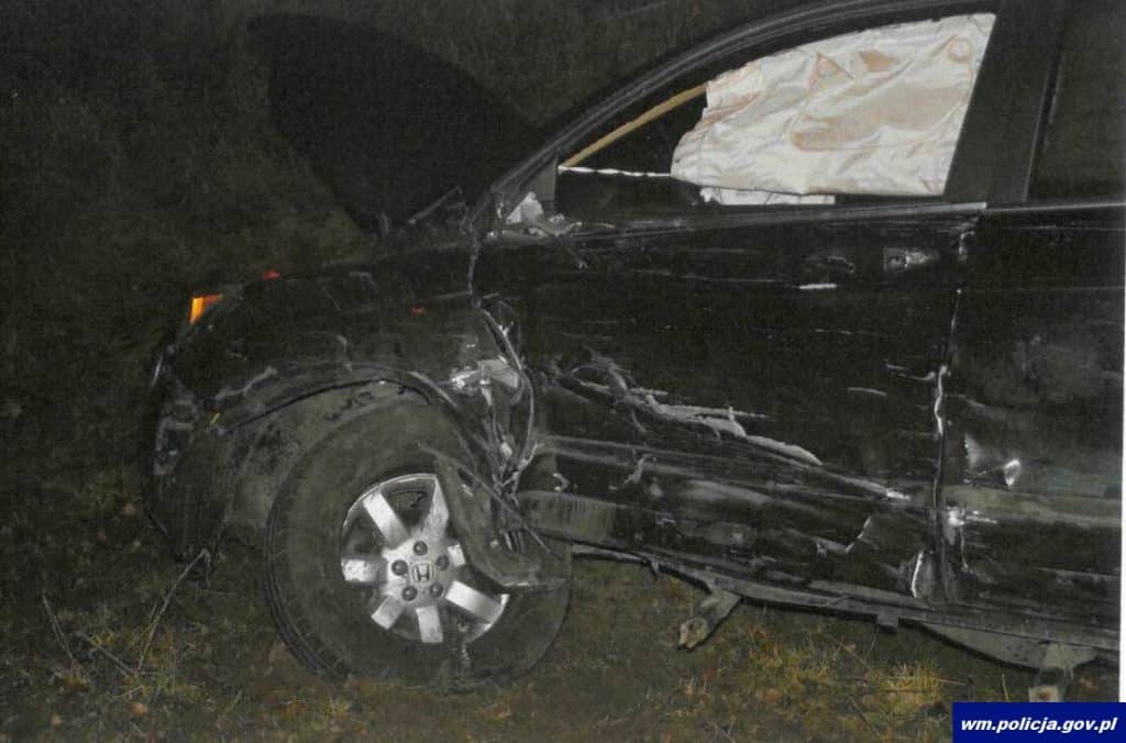 Uszkodzony czarny samoch&oacute;d po wypadku na drodze krajowej, widoczne zniszczenia po zderzeniu.