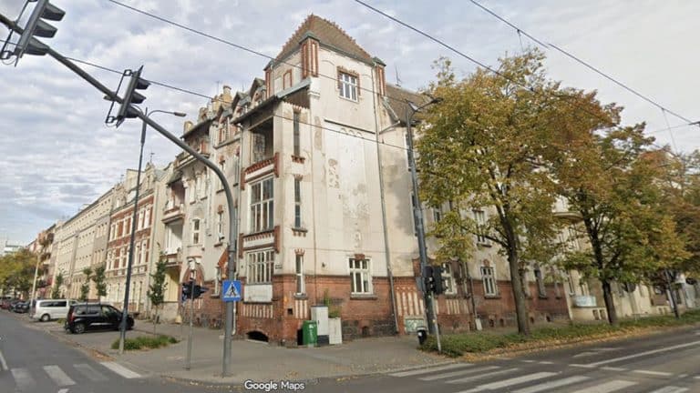 Zabytkowa kamienica w centrum Olsztyna przy ul. Mickiewicza 27, otoczona drzewami i ulicą z samochodami.