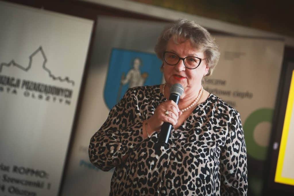 Kobieta w okularach i bluzce w leopardy wz&oacute;r m&oacute;wi do mikrofonu podczas wydarzenia.