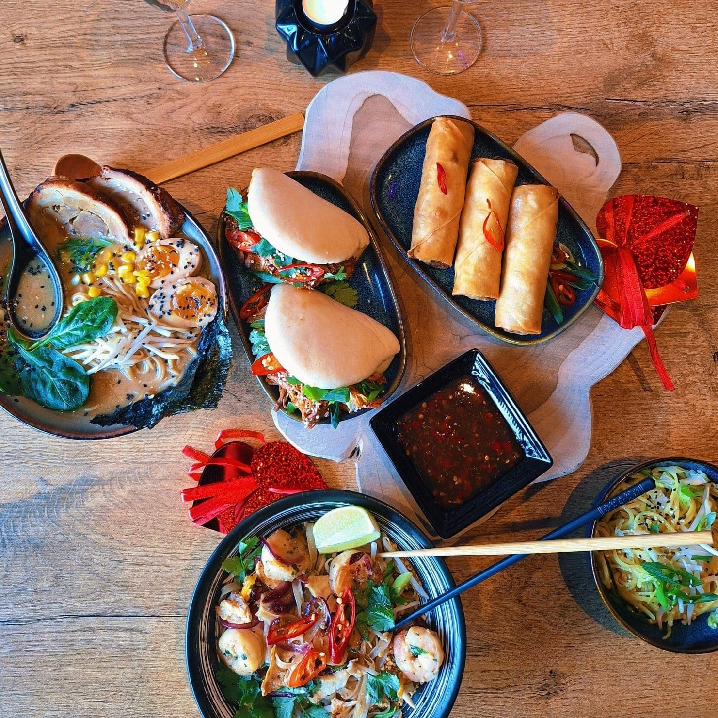 Zdjęcie r&oacute;żnorodnych dań azjatyckich na drewnianym stole, w tym spring rollsy, zupy i sałatki.