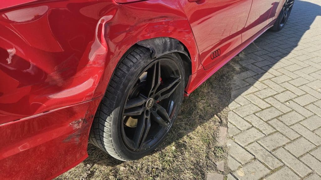 Uszkodzony czerwony samoch&oacute;d na rondzie, widoczny wywr&oacute;cony bok i uszkodzenia karoserii.