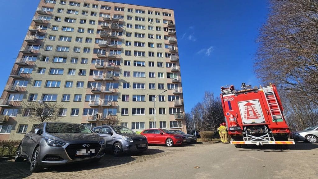 Wysoki blok mieszkalny z pojazdami służb ratunkowych przed budynkiem, na osiedlu w Olsztynie.