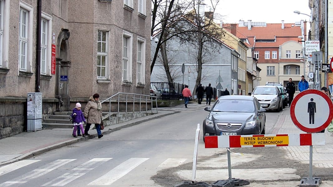 Ulica w centrum Olsztyna z zamkniętym placem budowy i samochodami na poboczu.