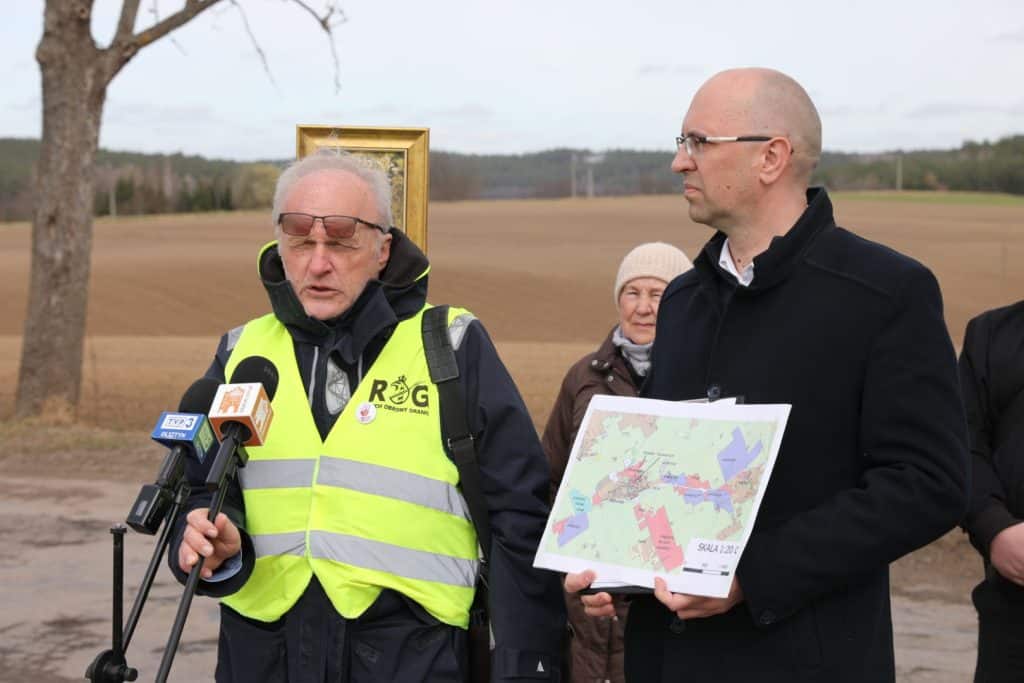 Mężczyzna w kamizelce odblaskowej i drugi z mapą na tle pola, podczas protestu w Gietrzwałdzie.