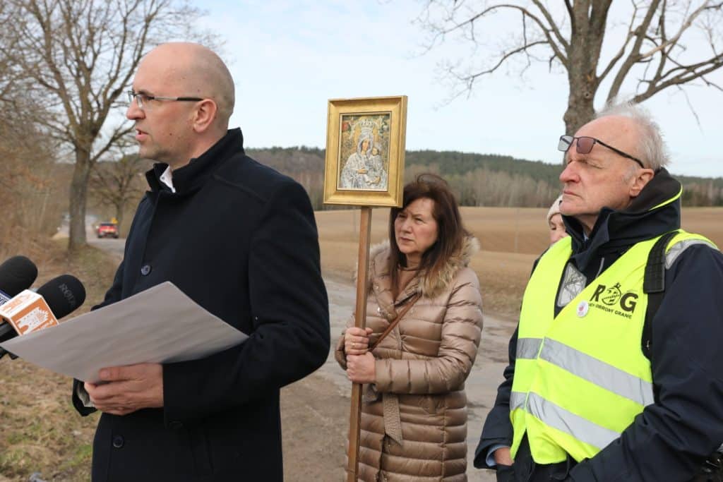 Grupa ludzi na tle pola, jedna osoba trzyma ikonę, inni słuchają podczas protestu.