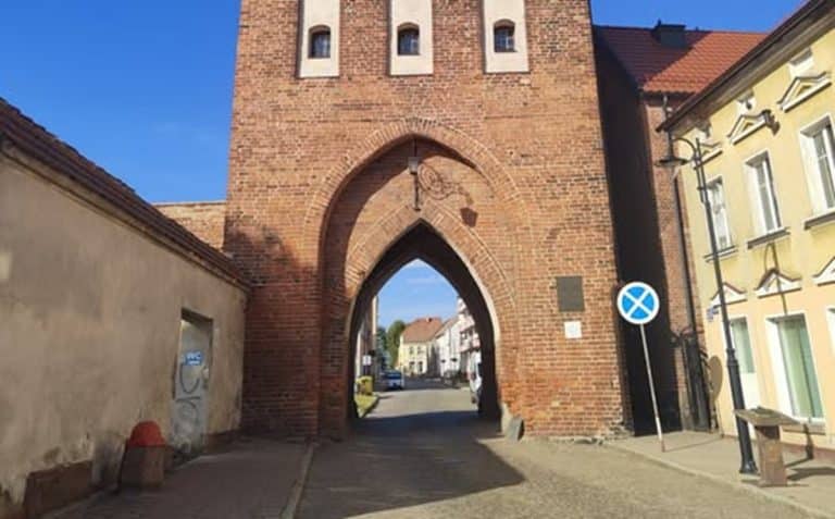 Widok średniowiecznej Bramy Kamiennej w Pasłęku, zbudowanej z czerwonej cegły, na tle nieba i okolicznych budynk&oacute;w.