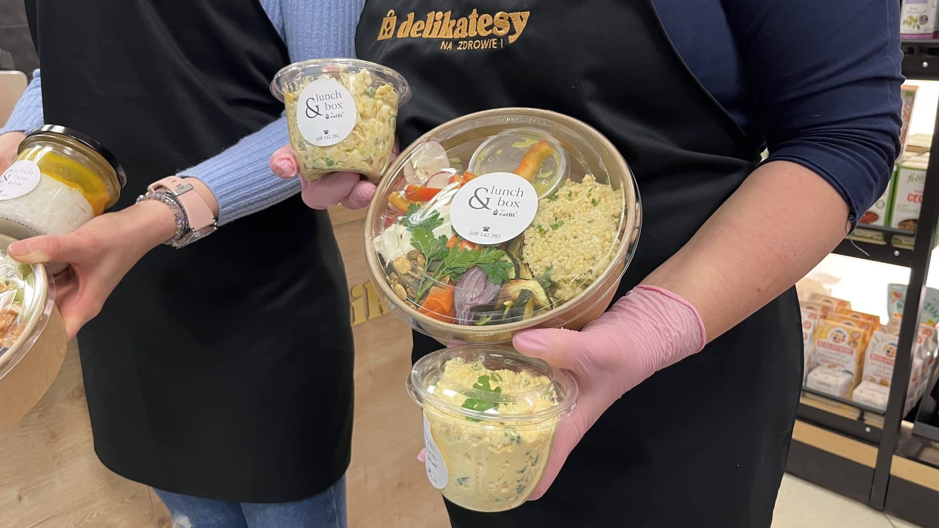 Dwie osoby trzymają r&oacute;żne pudełka z zdrowymi lunchami i sałatkami, w tle sklep spożywczy.