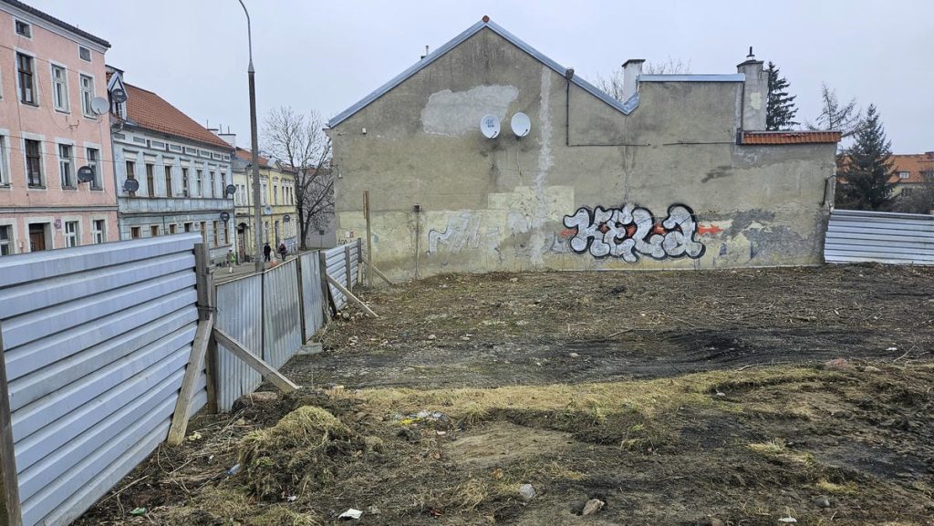 Teren przy szpitalu w Olsztynie przygotowany pod budowę nowoczesnego, przeszklonego budynku, z ogrodzeniem i graffiti na ścianie.