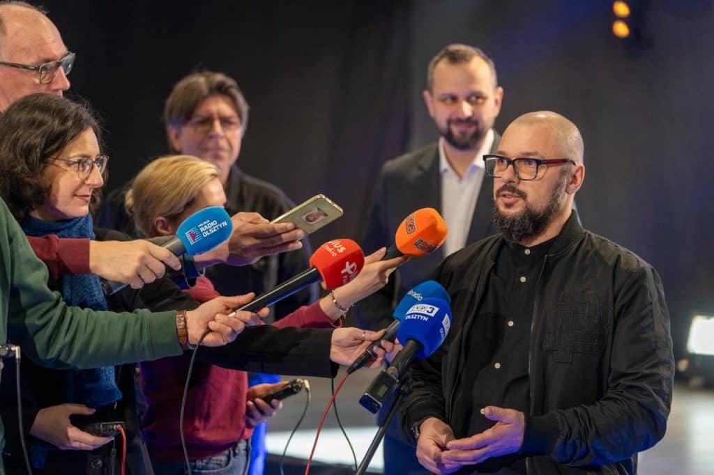 Mężczyzna w okularach i czarnej koszuli udziela wywiadu, wok&oacute;ł niego dziennikarze z mikrofonami.